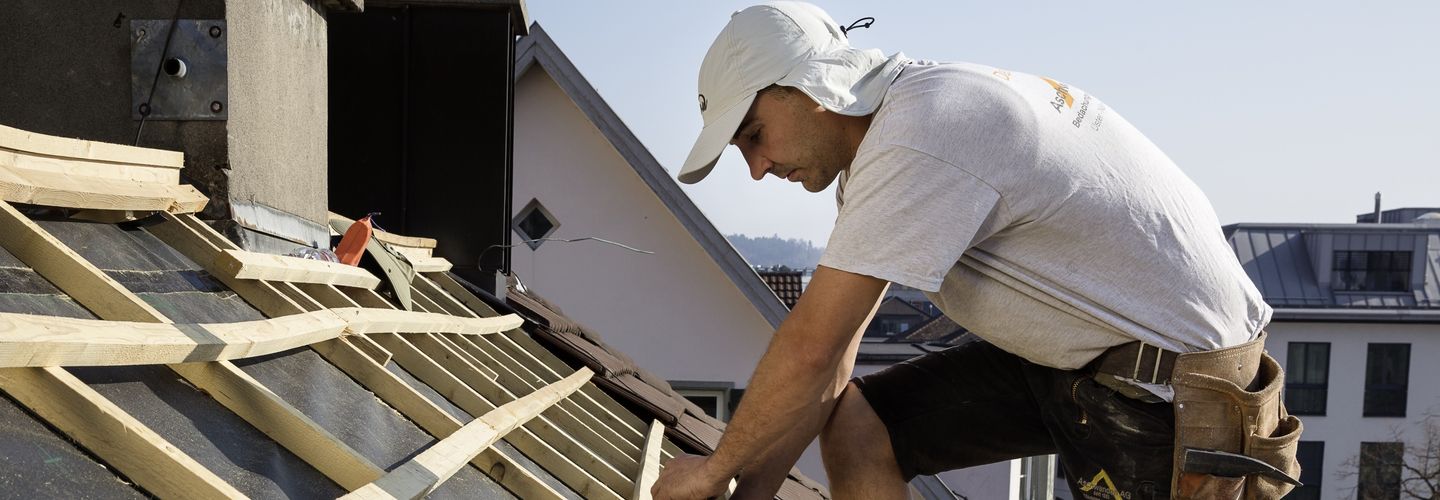 Dachdecker mit knielanger dunkler Hose, weissem T-Shirt und einer Kappe mit Stirnblende und Nackenschutz beim Anbringen von Dachlatten. 