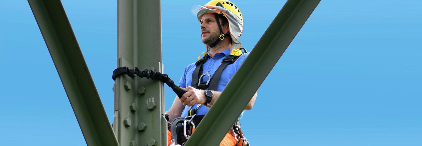 Un homme se tient en hauteur, attaché à un mât, portant sa protection solaire.