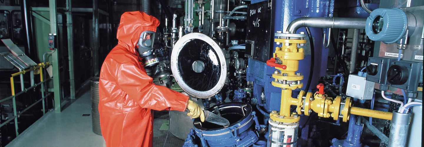 Une personne en tenue de protection contre les produits chimiques, avec un masque complet de protection respiratoire, prélève un échantillon dans un réservoir.