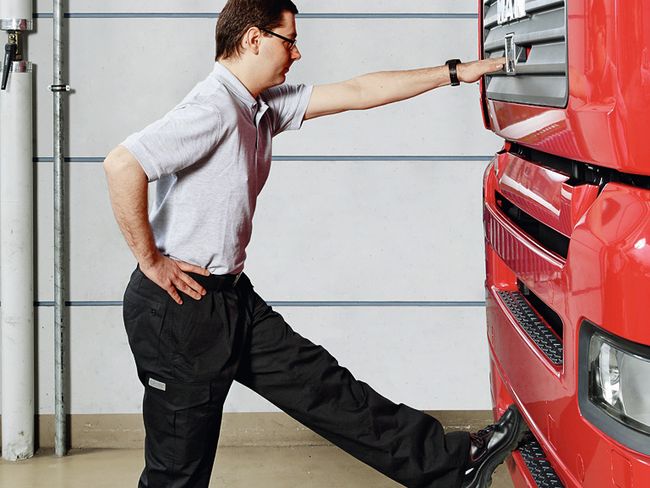 Ein Mann steht vor einem LKW. Er hebt das linke Bein an. Der Schuh ruht auf dem Kühlergrill, die Ferse senkt sich in Richtung Boden. Mit dem linken Arm stützt sich der Mann am Fahrzeug ab.