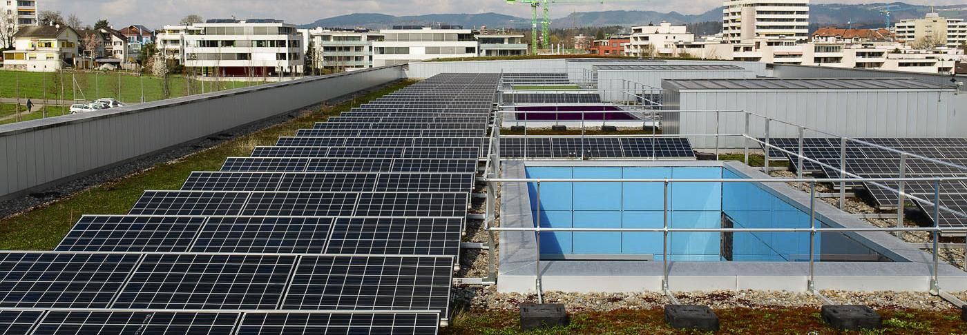 Aufnahme eines Flachdachs: Um den Dachrand zieht sich eine Brüstung. Links und rechts befinden sich je eine Reihe Solarmodule, in der Mitte Oblichter, die mit einem Metallgeländer gesichert sind. 