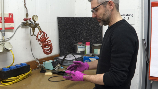 Un uomo dai capelli grigi con una t-shirt nera cambia i guanti da lavoro rosa a un banco da lavoro.