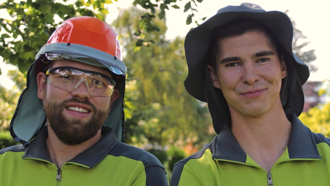 Fermo immagine dal video. Mezzo busto di Mario Enz (a sinistra) e Fabian Hodel (a destra), entrambi giardinieri paesaggisti campioni del mondo, che indossano un copricapo e indumenti da lavoro a maniche lunghe. Mario Enz indossa anche occhiali di protezione. Fabian Hodel fa il gesto «pollice su».