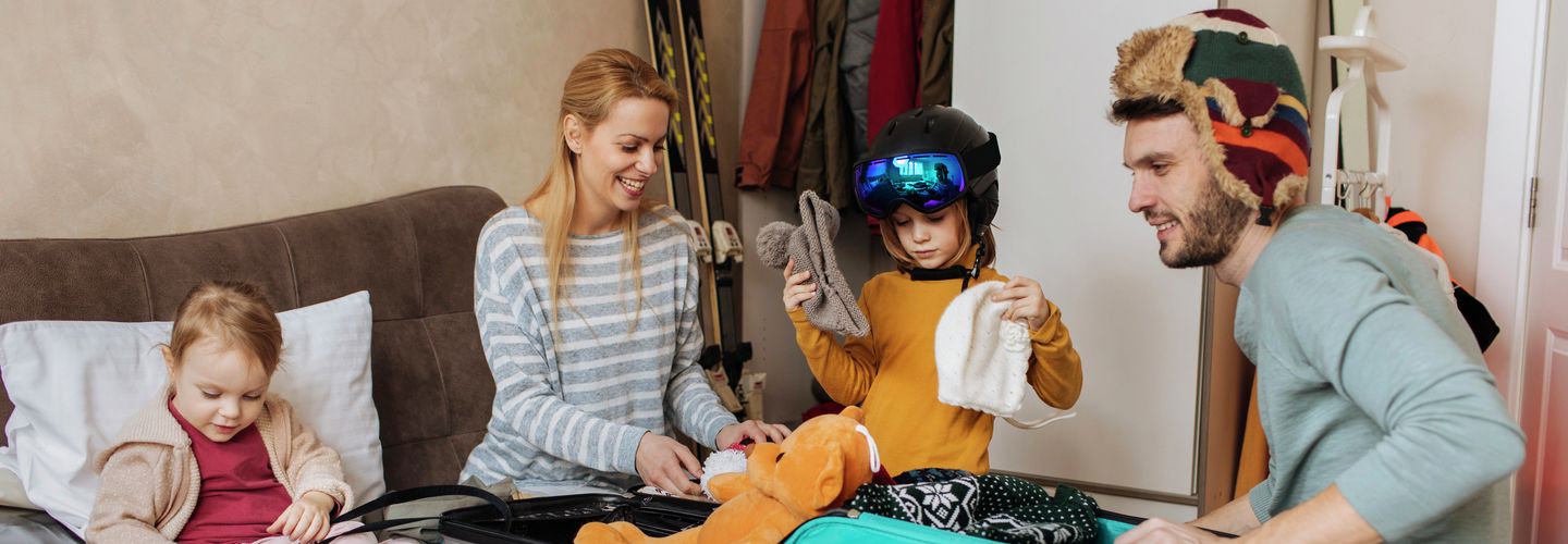 Eine fröhliche Familie packt lachend warme Kleidung in Koffer für ihren Winterurlaub.