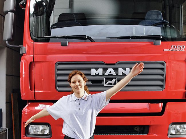 Eine Frau steht vor einem LKW. Sie hat die Arme waagrecht ausgestreckt und schwingt sie locker.