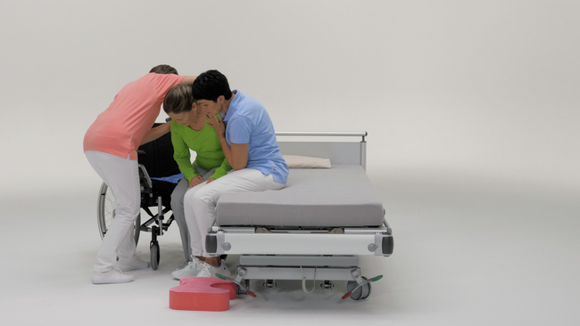 Wir sehen eine Frau, die auf der Kante eines Pflegebetts sitzt. Zwei unterstützende Personen helfen ihr dabei, sich in einen Rollstuhl zu setzen. Dabei positionieren sie ein Rutschbrett unter dem Becken der Frau. In kleinen Schritten und mit einer wiederholten Gewichtsverlagerung rutscht die Frau in Richtung Rollstuhl. Mit einem Footstool kann die Position der Füsse im Rollstuhl verbessert werden, oder die unterstützenden Personen nutzen einen Footstool, wenn sie in den Kniestand gehen.