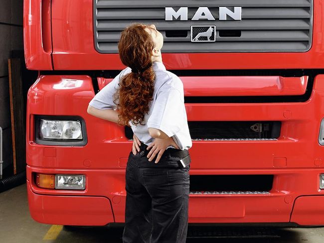Une femme se tient devant un camion. Ses mains se trouvent au niveau des reins. Son buste et sa tête sont légèrement inclinés vers l’arrière. Les coudes et les épaules sont enroulés vers la colonne vertébrale. Dans le même temps, elle pousse ses hanches vers l’avant.