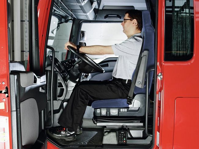 Un homme est assis au volant d’un camion. Il se tient droit et sa nuque repose contre l’appuie-tête. Les armes détendus reposent sur le volant, les épaules sont basses.