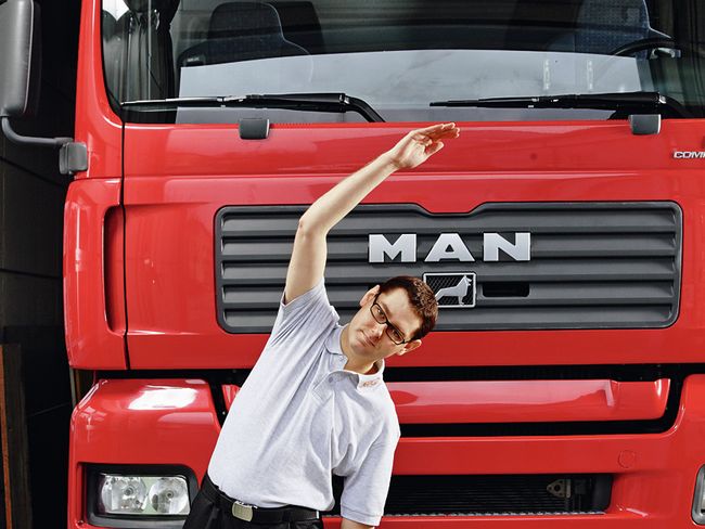 Ein Mann steht vor einem LKW. Er hat den rechten Arm angehoben und beugt den Oberkörper auf die linke Seite. Der linke Arm gleitet entlang des Oberschenkels in Richtung Knie.