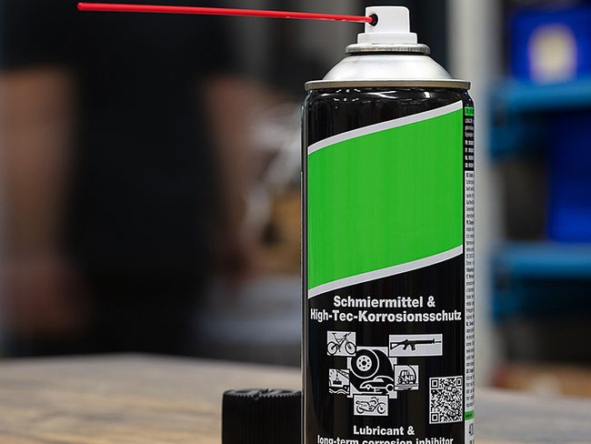 Zu sehen ist eine Spraydose mit schwarzer Grundierung, über die sich ein grüner Streifen zieht. Unter einer weissen Produktbezeichnung "Schmiermittel & High Tec-Korrosionsschutz" sind weisse Piktogramme für Einsatzgebiete wie Auto, Velo oder Seilbahn angebracht. 