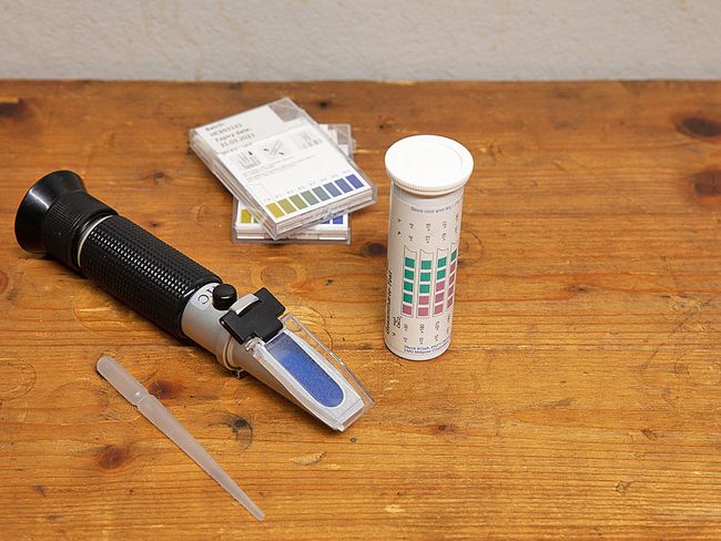 Les éléments suivants sont alignés sur une table en bois: une pipette en plastique, un réfractomètre, un tube à essai et, derrière, deux paquets de bandelettes de test.
