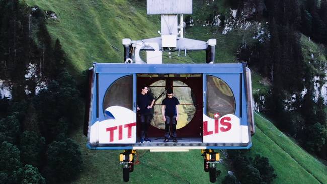 Fermo immagine dal video. Una funivia si libra sopra un precipizio. La porta è spalancata. Si vedono due giovani uomini, uno dei quali guarda verso l’abisso.