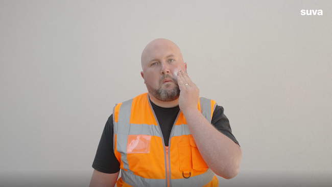 Bauarbeiter in orange-grau gestreifter Weste cremt sein Gesicht mit Sonnencreme ein.