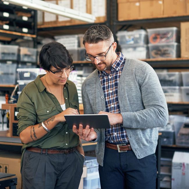 Eine Frau und ein Mann stehen in einer Werkstatt und schauen gemeinsam auf ein digitales Tablet, das der Mann hält. Die Frau zeigt auf das Display, während sie sich über die angezeigten Informationen austauschen.