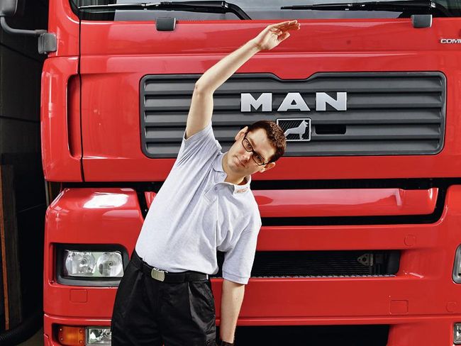 Un uomo è davanti a un camion. Ha sollevato il braccio destro e piega il busto sul lato sinistro. Il braccio sinistro svicola lungo la coscia verso il ginocchio.