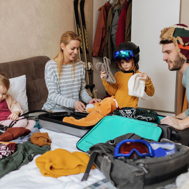 Eine fröhliche Familie packt lachend warme Kleidung in Koffer für ihren Winterurlaub.