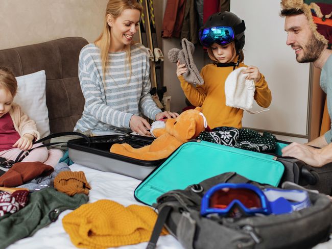 Eine fröhliche Familie packt lachend warme Kleidung in Koffer für ihren Winterurlaub.