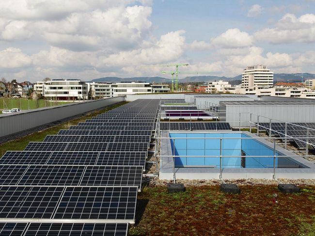 Aufnahme eines Flachdachs: Um den Dachrand zieht sich eine Brüstung. Links und rechts befinden sich je eine Reihe Solarmodule, in der Mitte Oblichter, die mit einem Metallgeländer gesichert sind. 