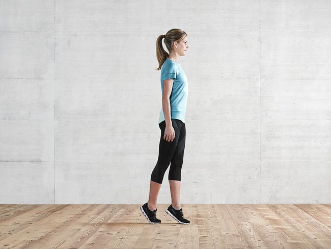 Eine Frau in Sportkleidung steht auf den Zehenspitzen. Ihr rechter Fuss steht dabei hinter dem linken Fuss.
